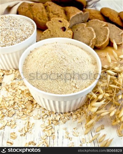 Flour oat and bran in two white bowls, oatmeal, oat stalks, bread and biscuits, cloth on a background of wooden boards