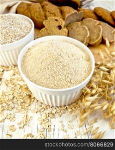 Flour oat and bran in two white bowls, oatmeal, oat stalks, bread and biscuits, cloth on a background of wooden boards