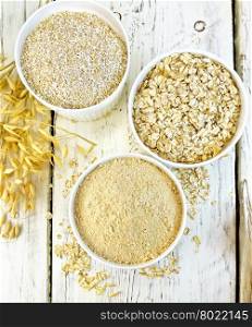 Flour oat, bran and oat flakes in three white bowls, stalks of oats on the background of the wooden planks on top