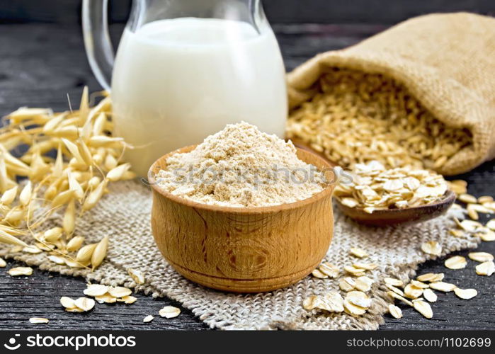 Flour oat in a bowl, milk in a jug, oatmeal in a spoon on burlap, grain in bag, oaten stalks on wooden board background