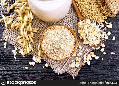 Flour oat in a bowl, milk in jug, oatmeal in spoon on burlap, grain in bag, oaten stalks on wooden board background from above