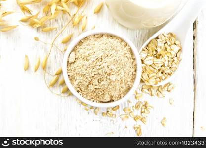 Flour oat in bowl, milk in a jug, oatmeal in spoon, oaten stalks on the background of white wooden board from above