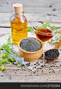 Flour of black caraway in a bowl, seeds in a spoon, oil in a bottle and sauceboat on burlap, sprigs of Nigella sativa with blue flowers and green leaves on wooden board