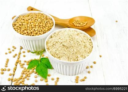 Flour soy and soybeans in two white bowls, spoons and green leaves on the background of light wooden boards