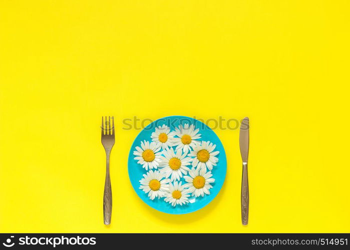 Flower chamomile daisy on blue plate and cutlery fork knife on yellow paper background Concept vegetarianism, healthy eating or diet Creative top view Copy space template for lettering text or your design.. Flower chamomile daisy on blue plate, cutlery fork knife on yellow paper background Concept vegetarianism, healthy eating or diet Creative top view Copy space template for lettering text or design