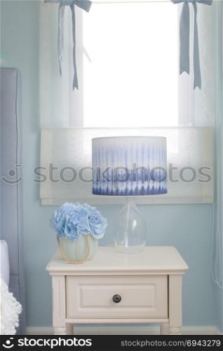 Flower jar and reading lamp on bedside table in light blue interior bedroom