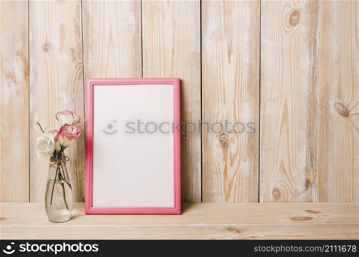 flower vase near white blank frame with pink border against wooden wall