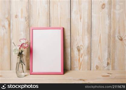 flower vase near white blank frame with pink border against wooden wall