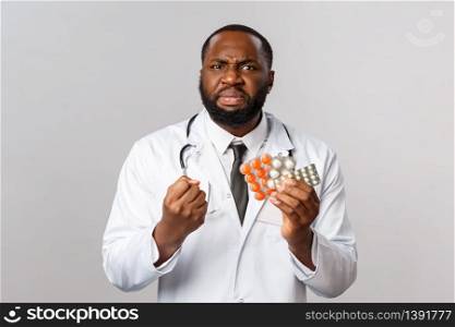 Flu, disease, healthcare and medicine concept. Angry, outraged african-american doctor furious over people selling drugs from coronavirus, shake fist and showing pills, dont approve scums.. Flu, disease, healthcare and medicine concept. Angry, outraged african-american doctor furious over people selling drugs from coronavirus, shake fist and showing pills, dont approve scums