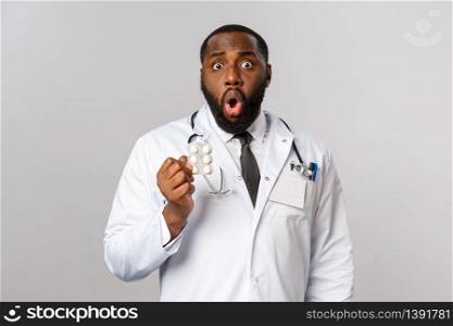 Flu, disease, healthcare and medicine concept. Shocked african-american doctor realise patient took pills wrong, didnt follow prescribtion, drop jaw gasping and holding medication, grey background.. Flu, disease, healthcare and medicine concept. Shocked african-american doctor realise patient took pills wrong, didnt follow prescribtion, drop jaw gasping and holding medication, grey background