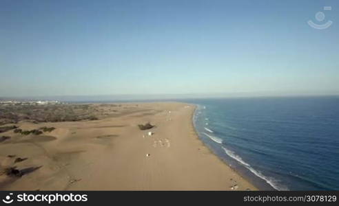 Flying over the coast with sand dunes and people relaxing by the clear blue ocean. Vacation and tourism on Gran Canaria, Canary Islands