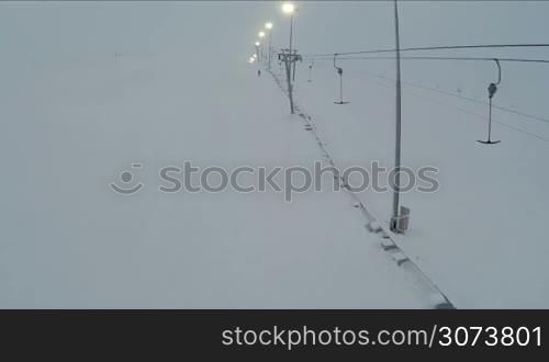 Flying over the working empty ski lift and lampposts along the hillside in the evening, few people skiing in the distance