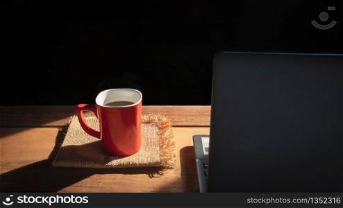 Focus at red coffee cup on napkins and blurred black laptop on wooden table with sunlight and shadow in black background, coffee break concept