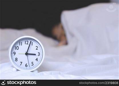 Focus at white round alarm clock in 3 o&rsquo;clock with blurred background of woman is sleeping on bed at night
