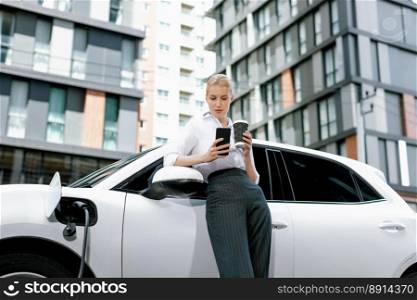 Focus businessman using phone, leaning on electric vehicle, holding coffee with blurred city residential condo buildings in background as progressive lifestyle by renewable and sustainable EV car.. Focus progressive woman using phone and holding coffee at charging station.