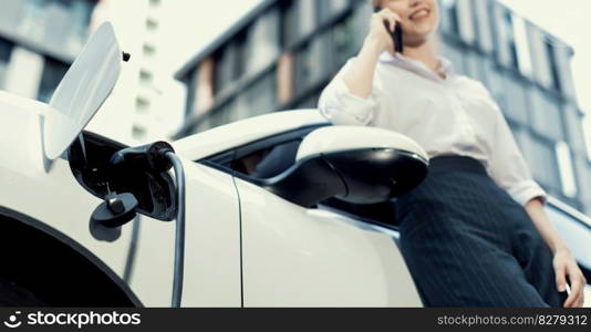 Focus charging-electric car with EV charger at charging station with blur businesswoman talking on phone with residential building in background as progressive lifestyle concept.. Focus electric car at charging station with blur background of progressive woman