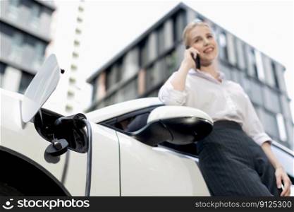 Focus charging-electric car with EV charger at charging station with blur businesswoman talking on phone with residential building in background as progressive lifestyle concept.. Focus electric car at charging station with blur background of progressive woman
