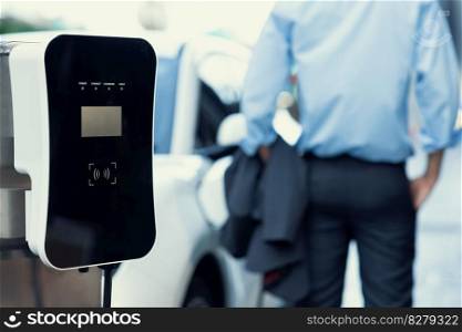 Focus closeup electric vehicle recharge battery at public charging station in the city area with blur businessman in background. EV car attached with electric charger for eco-friendly idea.. Focus closeup EV car recharge battery at public charging station.