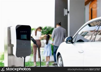 Focus closeup electric vehicle recharging battery from home electric charging station with blurred family in background. Renewable clean energy car for progressive eco awareness lifestyle concept.. Focus home charging station for EV car, blur progressive family in background.