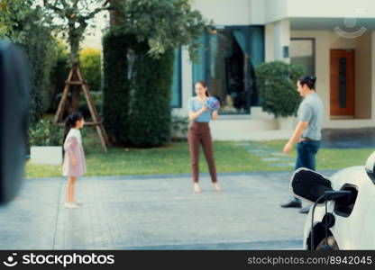 Focus closeup electric vehicle recharging battery from home electric charging station with blurred family in background. Renewable clean energy car for progressive eco awareness lifestyle concept.. Focus home charging station for EV car, blur progressive family in background.