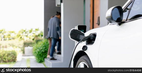 Focus closeup electric vehicle recharging battery from home electric charging station with blurred family in background. Renewable clean energy car for progressive eco awareness lifestyle concept.. Focus home charging station for EV car, blur progressive family in background.