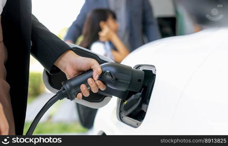 Focus closeup hand charging electric car, insert charger device into electric vehicle as progressive lifestyle concept of alternative green energy technology with blurred family in the background.. Focus hand charging progressive EV car with blurred family background.