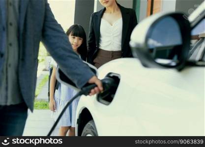 Focus closeup hand charging electric car, insert charger device into electric vehicle as progressive lifestyle concept of alternative green energy technology with blurred family in the background.. Focus hand charging progressive EV car with blurred family background.