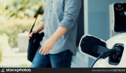 Focus electric car charging at home charging station with blurred progressive man walking in the background. Electric car using renewable clean for eco-friendly concept.. Focus EV charging station at home with blur progressive man in background.