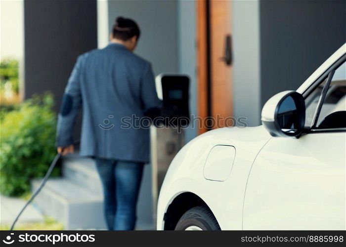Focus electric car charging at home charging station with blurred progressive man walking in the background. Electric car using renewable clean for eco-friendly concept.. Focus EV charging station at home with blur progressive man in background.