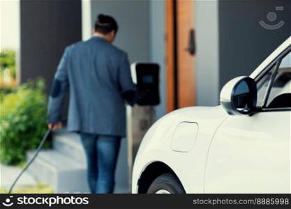 Focus electric car charging at home charging station with blurred progressive man walking in the background. Electric car using renewable clean for eco-friendly concept.. Focus EV charging station at home with blur progressive man in background.