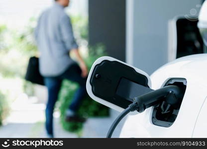 Focus electric car charging at home charging station with blurred progressive man walking in the background. Electric car using renewable clean for eco-friendly concept.. Focus EV charging station at home with blur progressive man in background.