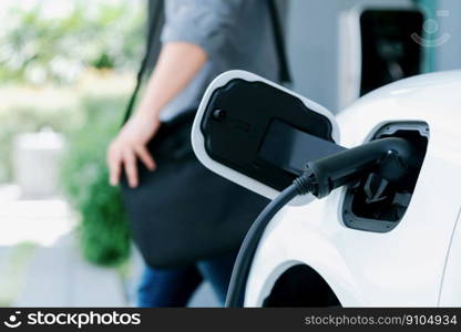 Focus electric car charging at home charging station with blurred progressive man walking in the background. Electric car using renewable clean for eco-friendly concept.. Focus EV charging station at home with blur progressive man in background.