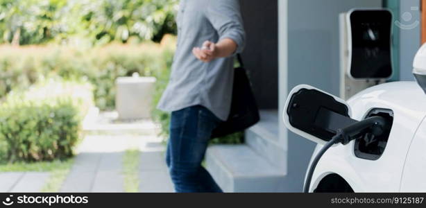 Focus electric car charging at home charging station with blurred progressive man walking in the background. Electric car using renewable clean for eco-friendly concept.. Focus EV charging station at home with blur progressive man in background.