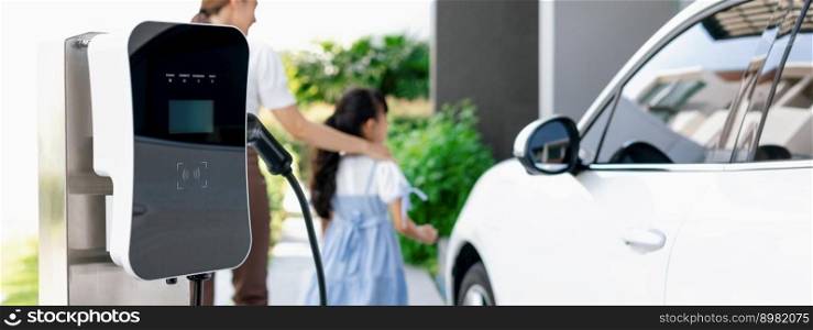 Focus electric charging station power by clean energy for electric vehicle at home with blurred progressive woman and girl walking in background. Home charging station for electric engine car concept. Focus progressive home EV charging point with blur woman and girl in background.