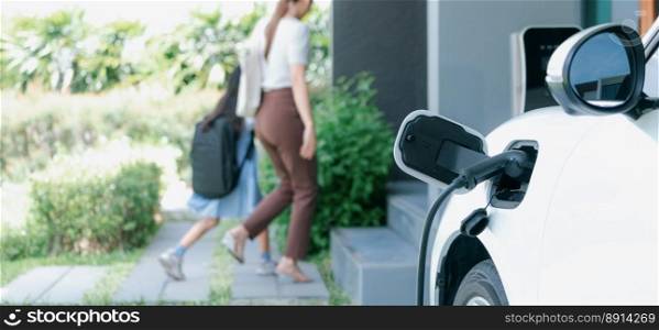 Focus electric vehicle recharging at home charging station plugged in with EV charger device with blurred background of progressive mother and daughter walking as concept for sustainability of energy.. Focus EV car charging at home charging point with blurred background of family.