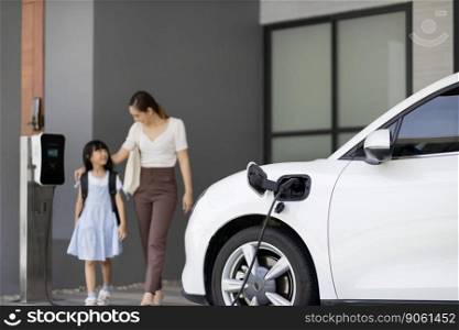 Focus EV car recharging at home charging station with blurred progressive woman and young girl in background for alternative clean energy technology concept for renewable electric vehicle.. Focus progressive EV car at home with blur woman in background.