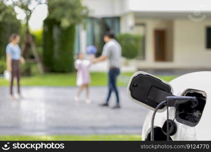 Focus EV charger connected with EV car at outdoor home charging station with blur progressive family playing together in the background. EV car for alternative clean energy concept.. Focus EV charger recharge EV car at home with progressive family in background.