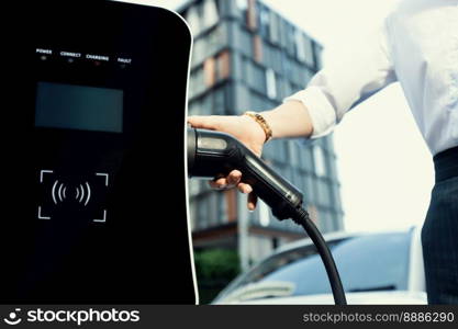 Focus EV charger plug and electric car at public charging station with blur progressive businesswoman holding charger and apartment condo building in background. Eco friendly electric vehicle concept.. Focus EV charger plug with blur progressive woman charge EV car in background.