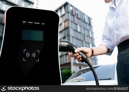 Focus EV charger plug and electric car at public charging station with blur progressive businesswoman holding charger and apartment condo building in background. Eco friendly electric vehicle concept.. Focus EV charger plug with blur progressive woman charge EV car in background.