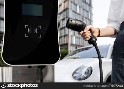 Focus EV charger plug and electric car at public charging station with blur progressive businesswoman holding charger and apartment condo building in background. Eco friendly electric vehicle concept.. Focus EV charger plug with blur progressive woman charge EV car in background.