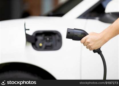 Focus hand holding EV charger plug with blurred background of progressive electric vehicle and socket parking in home garage with electric charging station powered by clean and sustainable energy.. Focus hand hold EV plug with blur image of progressive car at charging station.