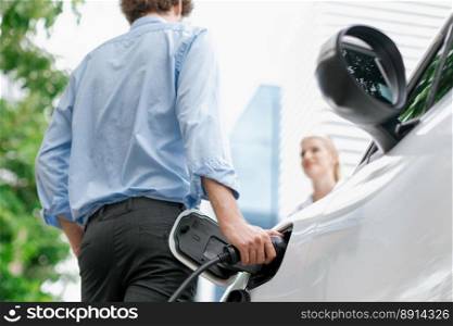 Focus hand insert EV charger to electric vehicle at public charging point in car park with blur business people in backdrop, eco-friendly lifestyle by rechargeable car for progressive concept.. Focus hand insert progressive EV charger with blurred background.