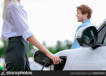 Focus hand insert EV charger to electric vehicle at public charging point in car park with blur business people in backdrop, eco-friendly lifestyle by rechargeable car for progressive concept.. Focus hand insert progressive EV charger with blurred background.