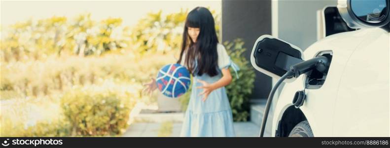 Focus home electric charging station for electric vehicle as alternative clean sustainable energy technology concept with blurred progressive young girl playing in the background at her home.. Progressive technology concept of focus EV charger with blur young girl backdrop