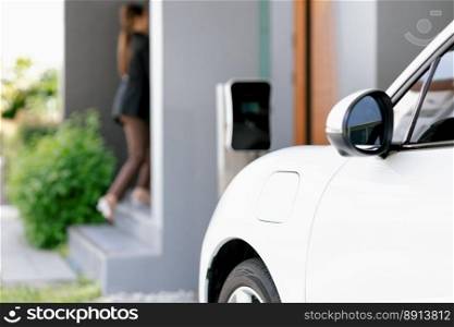 Focus image of electric vehicle recharging battery at home charging station with blurred woman walking in the background. Progressive concept of green energy technology applied in daily lifestyle.. Focus EV car at home charging station with blur progressive woman in the back.