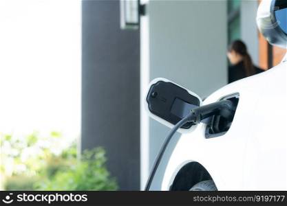 Focus image of electric vehicle recharging battery at home charging station with blurred woman walking in the background. Progressive concept of green energy technology applied in daily lifestyle.. Focus EV car at home charging station with blur progressive woman in the back.