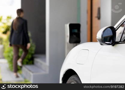 Focus image of electric vehicle recharging battery at home charging station with blurred woman walking in the background. Progressive concept of green energy technology applied in daily lifestyle.. Focus EV car at home charging station with blur progressive woman in the back.