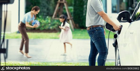 Focus image of progressive man charging electric car from home charging station with blur mother and daughter playing together in the background.. Focus image of progressive man charging EV car with his blur family background.