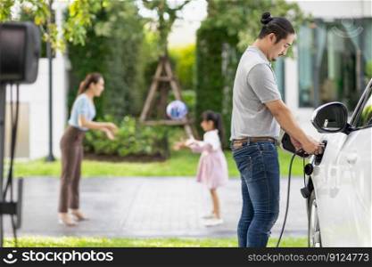 Focus image of progressive man charging electric car from home charging station with blur mother and daughter playing together in the background.. Focus image of progressive man charging EV car with his blur family background.