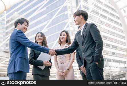 Focus on business men wearing formal suit and shake hands to deal agreement outside with blur background of colleagues